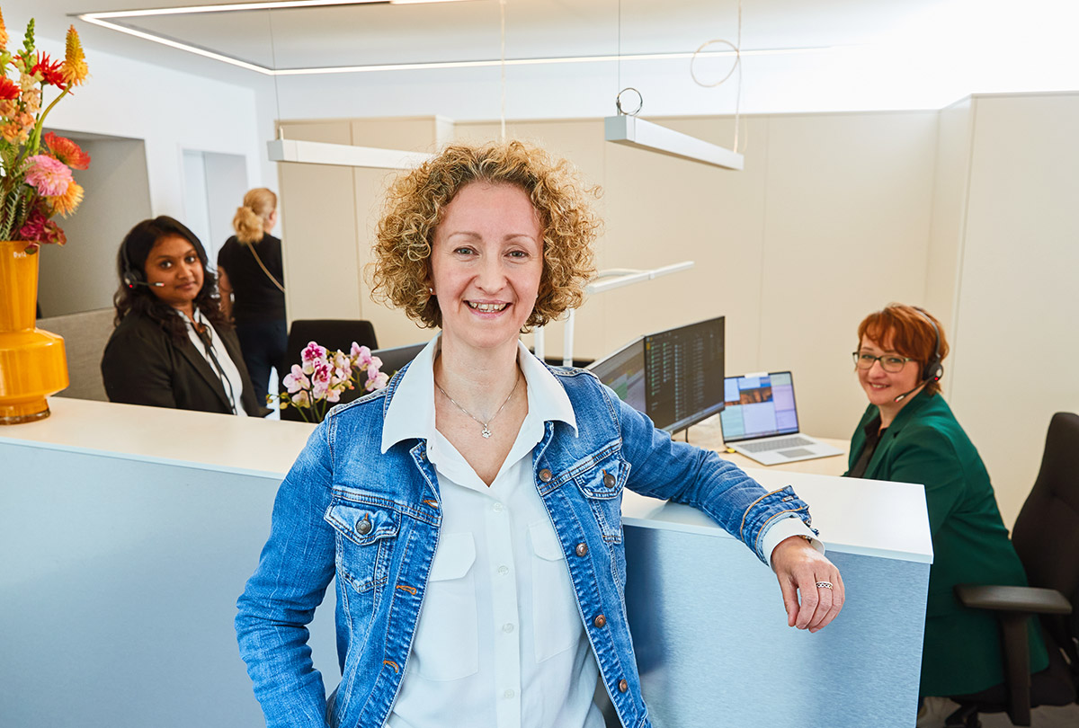 Frau mit lockigem Haar und Jeansjacke steht in einem modernen Büro, im Hintergrund sitzen zwei Personen an Computern, weitere Personen stehen im Raum, Kontaktaufnahme bei Global Westfalen