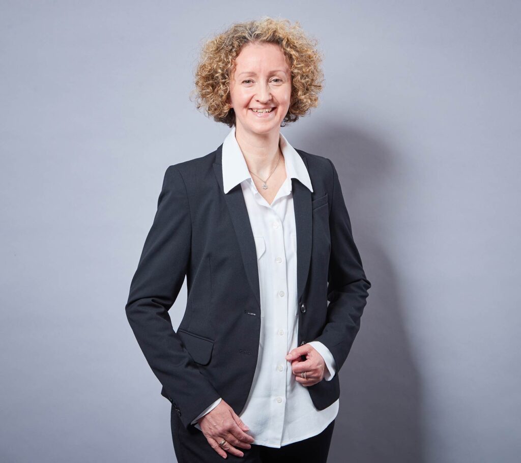 Sabine Linnenberg steht vor grauem Hintergrund, trägt schwarzen Blazer und weiße Bluse, hat lockige blonde Haare